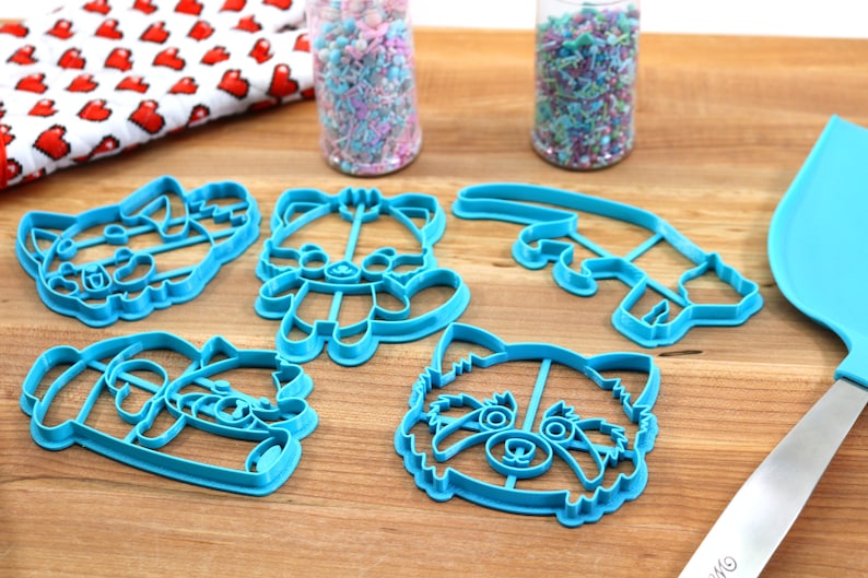 May include: Five blue plastic cookie cutters shaped like red pandas. The cutters are on a wooden surface with two jars of sprinkles in the background.