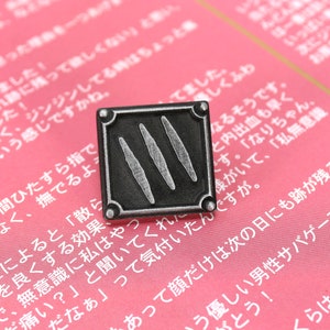 以下が含まれることがあります： 正方形のデザインで、3本の縦線が描かれた銀色の金属製のピン。ピンはピンクの表面に置かれ、日本語のテキストが書かれている。