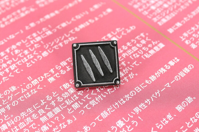 以下が含まれることがあります： 正方形のデザインで、3本の縦線が描かれた銀色の金属製のピン。ピンはピンクの表面に置かれ、日本語のテキストが書かれている。