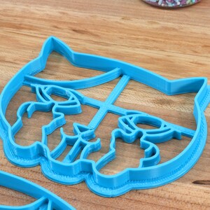 May include: A blue plastic cookie cutter shaped like a cat's face. The cutter has a detailed design with a cat's eyes, nose, and whiskers.