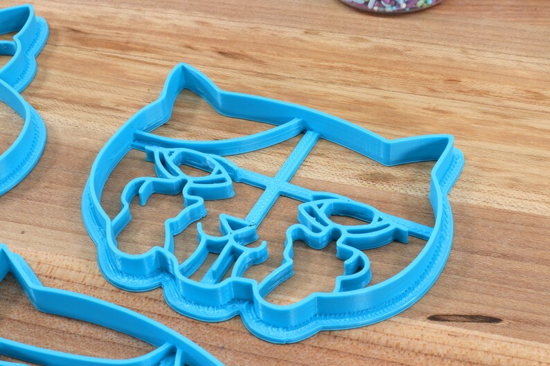 May include: A blue plastic cookie cutter shaped like a cat's face. The cutter has a detailed design with a cat's eyes, nose, and whiskers.
