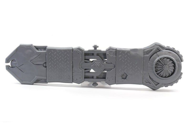 May include: A gray plastic toy with a detailed design, featuring a fan blade and a series of interlocking gears. The toy is designed to resemble a futuristic weapon or tool.