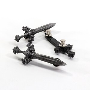 May include: Three silver-toned metal pins in the shape of swords and a cross. The swords have intricate details and the cross is simple. The pins are on a white background.