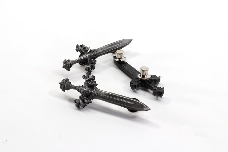 May include: Three silver-toned metal pins in the shape of swords and a cross. The swords have intricate details and the cross is simple. The pins are on a white background.