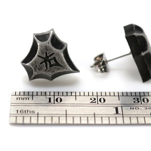 May include: A pair of silver stud earrings with a textured, geometric design. One earring features a Japanese character, "拓", in black. The earrings are shown next to a ruler, measuring approximately 1 cm in diameter.