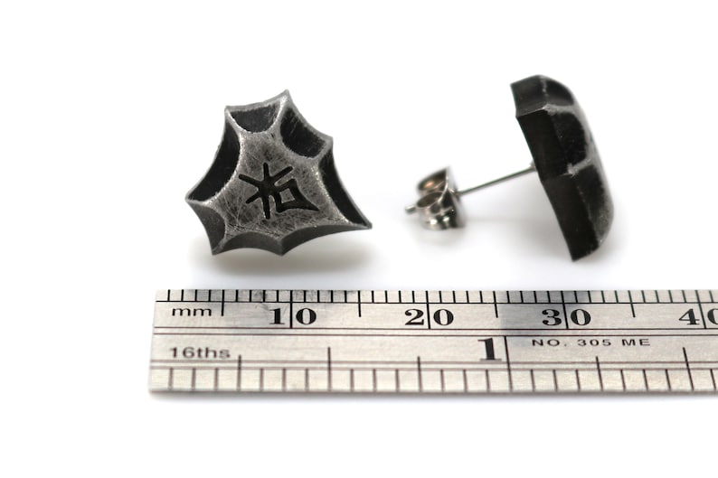 May include: A pair of silver stud earrings with a textured, geometric design. One earring features a Japanese character, "拓", in black. The earrings are shown next to a ruler, measuring approximately 1 cm in diameter.