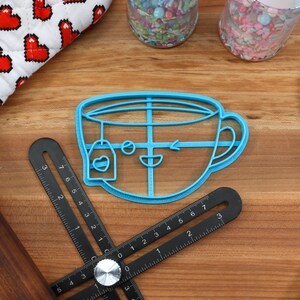 May include: A blue plastic cookie cutter shaped like a teacup with a teabag hanging from the rim. The teacup has a smiling face with a winking eye and a small arrow drawn on the side.