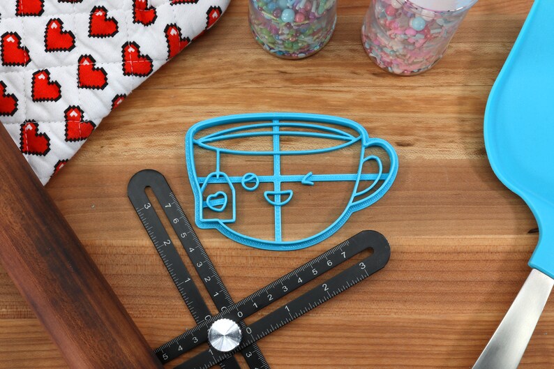 May include: A blue plastic cookie cutter shaped like a teacup with a teabag hanging from the rim. The teacup has a smiling face with a winking eye and a small arrow drawn on the side.