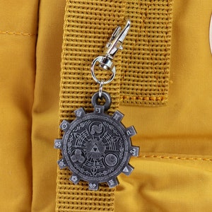 May include: A silver-colored metal charm with a detailed design of a circular symbol with a triangular shape in the center. The charm is attached to a silver-colored metal chain and is hanging from a yellow backpack.