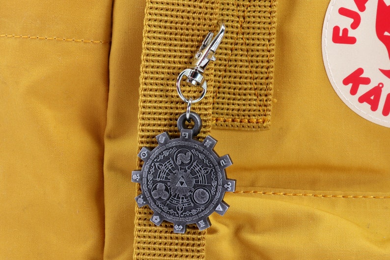 May include: A silver-colored metal charm with a detailed design of a circular symbol with a triangular shape in the center. The charm is attached to a silver-colored metal chain and is hanging from a yellow backpack.