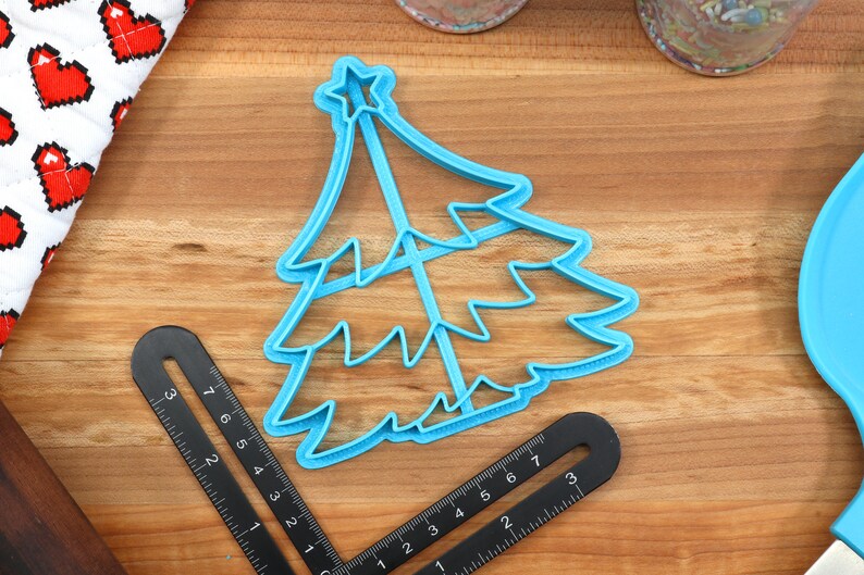 Op de afbeelding: Een blauwe plastic kerstboomkoekjesvormer met een ster bovenop. De vormer ligt op een houten oppervlak.