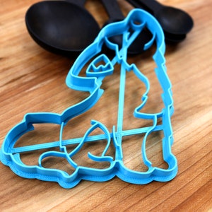 May include: A blue plastic cookie cutter shaped like a dog sitting down. The cutter is on a wooden surface.