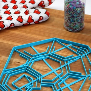 May include: A blue plastic snowflake cookie cutter with multiple layers of intricate designs. The cutter is on a wooden surface with two jars of sprinkles in the background.