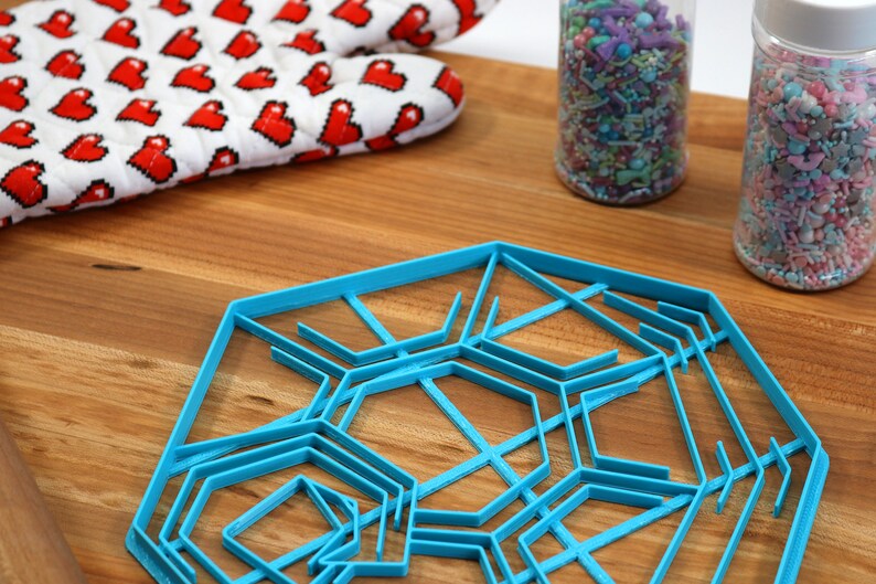 May include: A blue plastic snowflake cookie cutter with multiple layers of intricate designs. The cutter is on a wooden surface with two jars of sprinkles in the background.