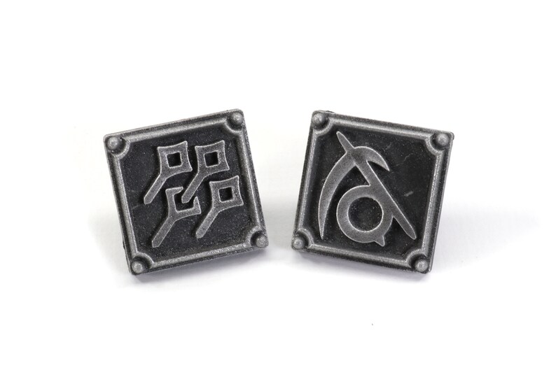 May include: Two silver-toned metal pins with square designs. One pin features a stylized symbol of a scythe and the other pin features a stylized symbol of four squares.