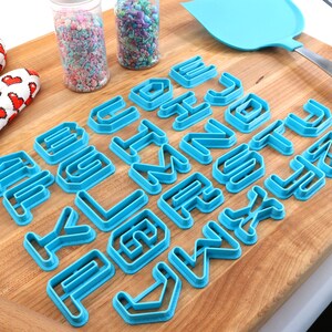 May include: A set of blue plastic alphabet cookie cutters on a wooden cutting board. The cutters are arranged in alphabetical order. There are two jars of sprinkles in the background.