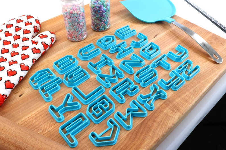 May include: A set of blue plastic alphabet cookie cutters on a wooden cutting board. The cutters are arranged in alphabetical order. There are two jars of sprinkles in the background.