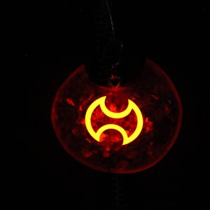 May include: A red pendant with a yellow symbol in the shape of a stylized crescent moon. The pendant is made of resin and has a glittery finish.