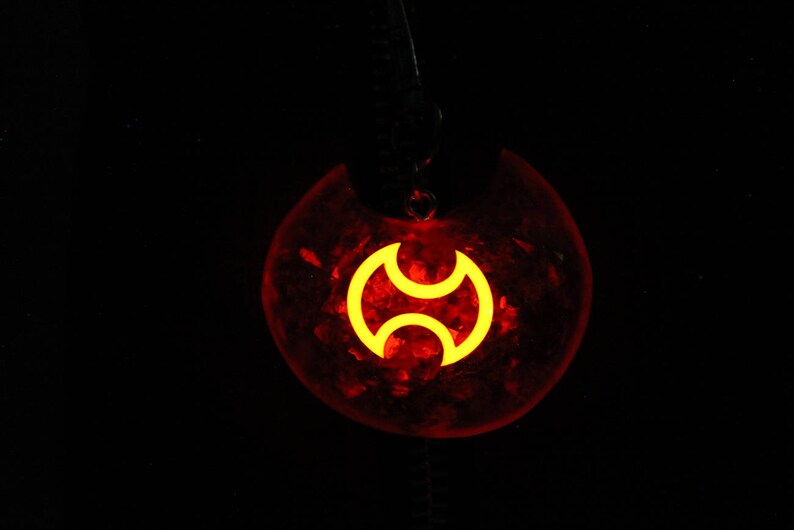 May include: A red pendant with a yellow symbol in the shape of a stylized crescent moon. The pendant is made of resin and has a glittery finish.