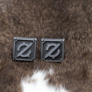 May include: A pair of silver cufflinks with a stylized letter 'Z' design. The cufflinks are square shaped and have a textured surface.