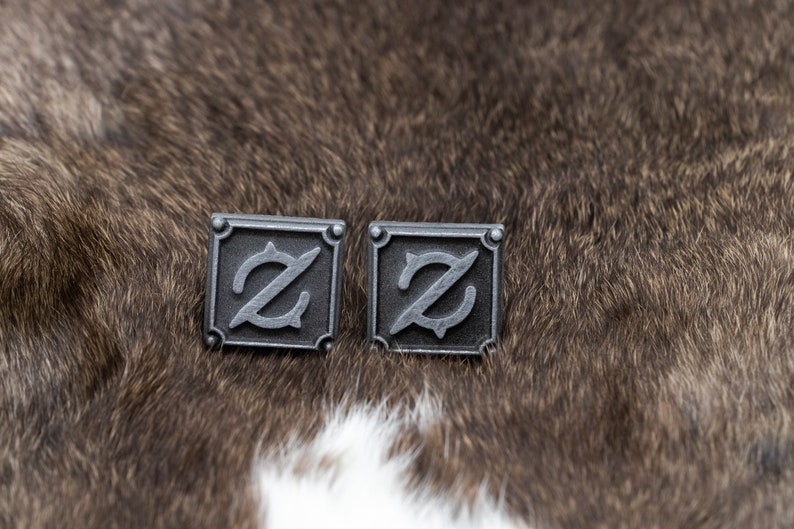 May include: A pair of silver cufflinks with a stylized letter 'Z' design. The cufflinks are square shaped and have a textured surface.