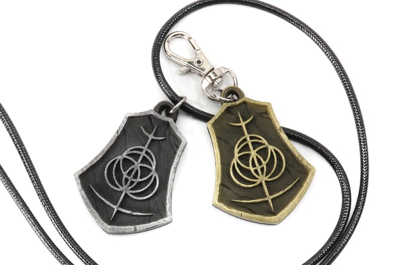 May include: Two silver and gold pendants with a black cord necklace. The pendants are shaped like shields with a circular design and a vertical line in the center.