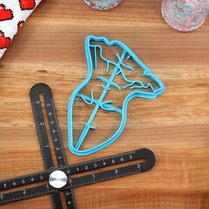 May include: A blue plastic cookie cutter shaped like a Christmas tree with a star on top. The cutter is on a wooden surface.