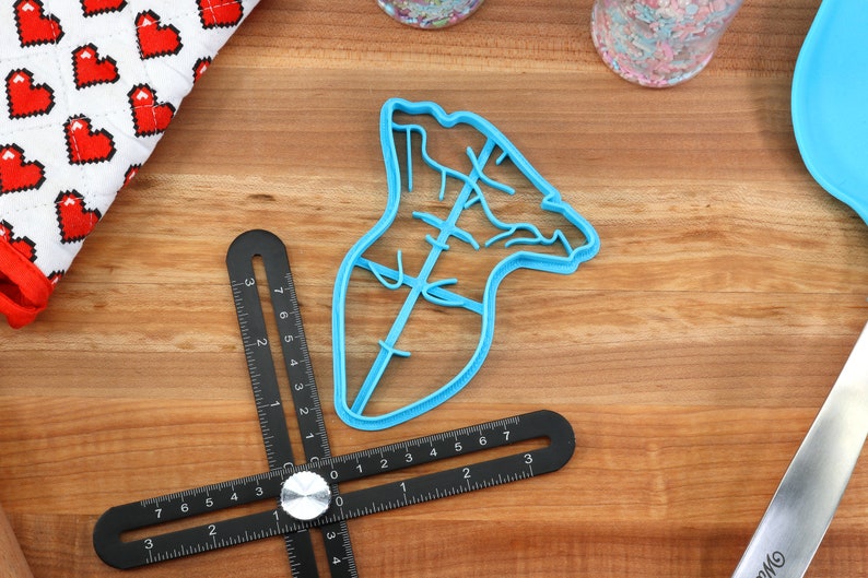May include: A blue plastic cookie cutter shaped like a Christmas tree with a star on top. The cutter is on a wooden surface.