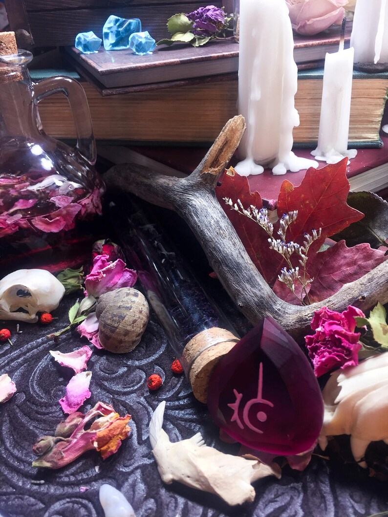 Op de afbeelding: Een donkerpaarse glazen fles met een kurken stop, een kleine schedel, een houten geweistuk, een paarse glazen edelsteen met een symbool en gedroogde bloemen op een zwarte stoffen achtergrond.