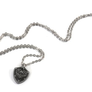 May include: A silver chain necklace with a pendant shaped like a shield with a design of a winged figure holding a sword.