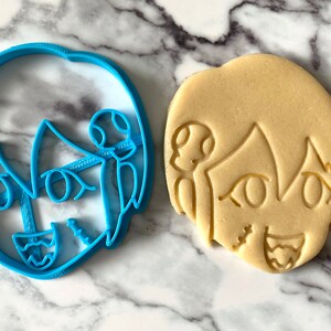 Pode incluir: Um cortador de biscoitos de pl&aacute;stico azul e um biscoito bege em forma de personagem de desenho animado com um sorriso largo e olhos grandes.
