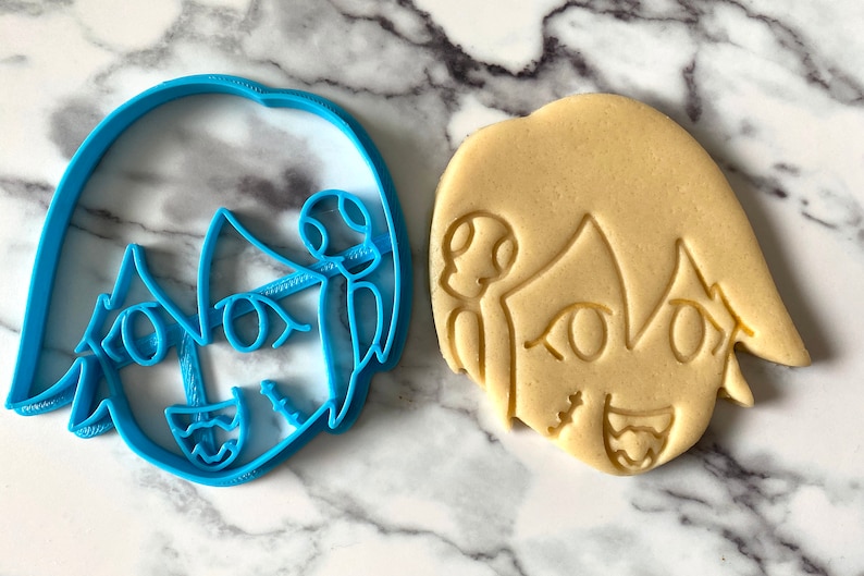 Pode incluir: Um cortador de biscoitos de pl&aacute;stico azul e um biscoito bege em forma de personagem de desenho animado com um sorriso largo e olhos grandes.
