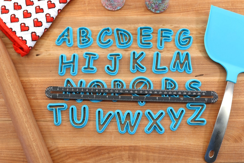 Op de afbeelding: Een set blauwe plastic alfabetkoekjesuitstekers op een houten oppervlak. De uitstekers zijn alfabetisch gerangschikt en worden omringd door een liniaal, een deegroller en een spatel.