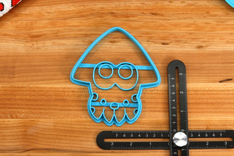 May include: A blue plastic cookie cutter shaped like a cartoon squid with a large head and a small body. The cutter has a detailed design with eyes, a mouth, and tentacles.