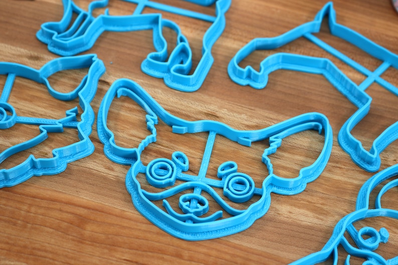 Puede incluir: Un juego de cortadores de galletas de pl&aacute;stico azul con forma de diferentes razas de perros. Los cortadores est&aacute;n sobre una superficie de madera.
