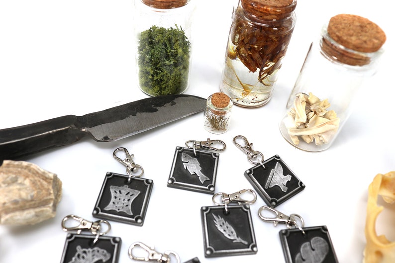 May include: A set of eight silver keychains with different symbols on black backgrounds. The keychains are arranged on a white surface with a knife, a small bottle of dried flowers, and three glass jars containing various items.