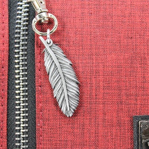 Op de afbeelding: Close-up van een rode, getextureerde stof met een zwarte rits en een zilveren veerketting. De bedel hangt aan een goudkleurige sluiting. De stof heeft een subtiel patroon.