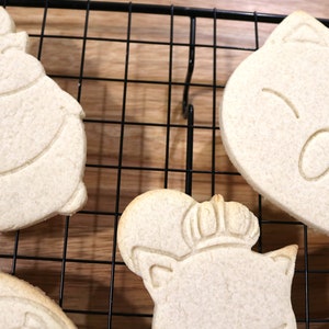 May include: Five plain sugar cookies on a black wire cooling rack. The cookies are shaped like cartoon characters with simple facial features.
