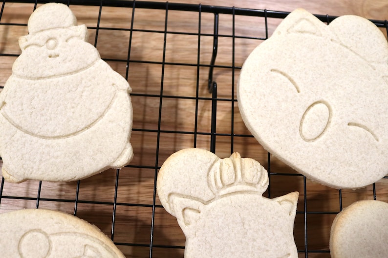 May include: Five plain sugar cookies on a black wire cooling rack. The cookies are shaped like cartoon characters with simple facial features.