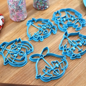 May include: Five blue plastic cookie cutters shaped like cartoon faces. The cutters are on a wooden surface.