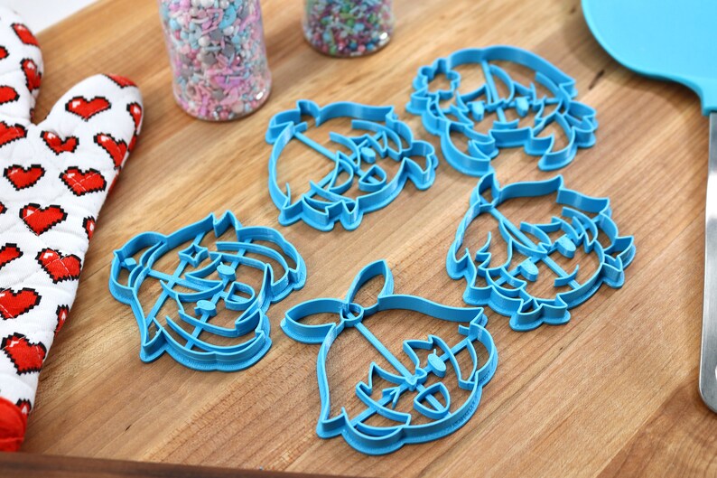 May include: Five blue plastic cookie cutters shaped like cartoon faces. The cutters are on a wooden surface.