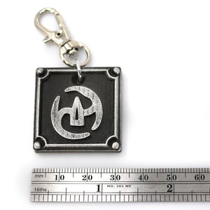 May include: A silver and black metal keychain with a stylised symbol on a square tag. The symbol is a stylised three-pronged design with a central point. The keychain has a silver metal clasp.