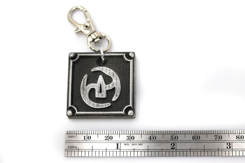 May include: A silver and black metal keychain with a stylised symbol on a square tag. The symbol is a stylised three-pronged design with a central point. The keychain has a silver metal clasp.