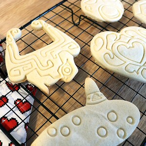 May include: A close-up of four cookies on a black wire rack. The cookies are shaped like a heart, a video game character, a musical instrument, and a heart with a design inside. The cookies are light brown in color and have a slightly crispy texture.