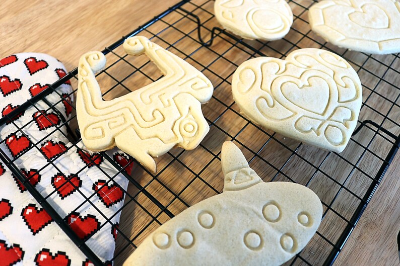 May include: A close-up of four cookies on a black wire rack. The cookies are shaped like a heart, a video game character, a musical instrument, and a heart with a design inside. The cookies are light brown in color and have a slightly crispy texture.