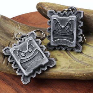 May include: Two silver-colored keychains shaped like a square with a frowning face. The keychains have a textured, metallic finish.