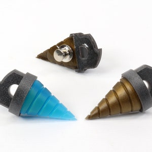 May include: Three drill-shaped pins with a unique design. One pin is blue, another is bronze, and the third is a combination of bronze and black. Each pin has a dark gray ring and a black backing with a pin clasp.