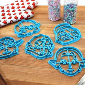 May include: Five blue plastic cookie cutters shaped like cartoon faces. The cutters are on a wooden surface with a red and white oven mitt, two jars of sprinkles, and a blue spatula.