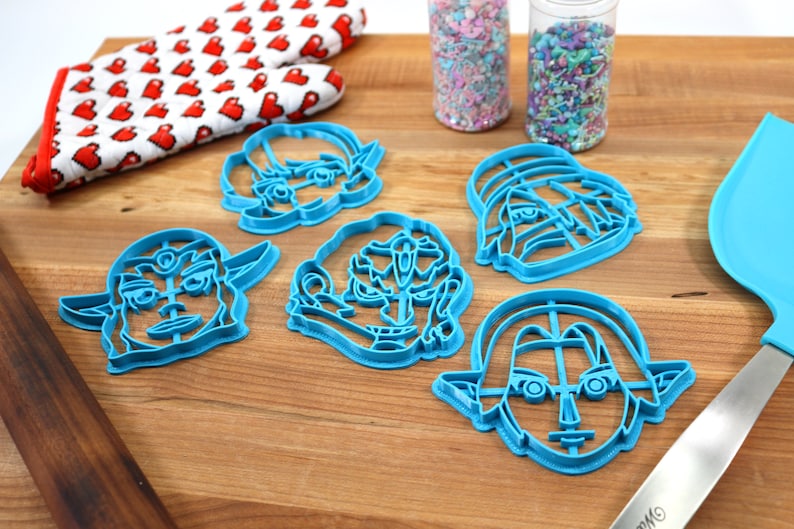 May include: Five blue plastic cookie cutters shaped like cartoon faces. The cutters are on a wooden surface with a red and white oven mitt, two jars of sprinkles, and a blue spatula.