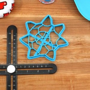 May include: A blue plastic cookie cutter shaped like a star with a geometric design. The cutter is about 3 inches in diameter and is sitting on a wooden surface.